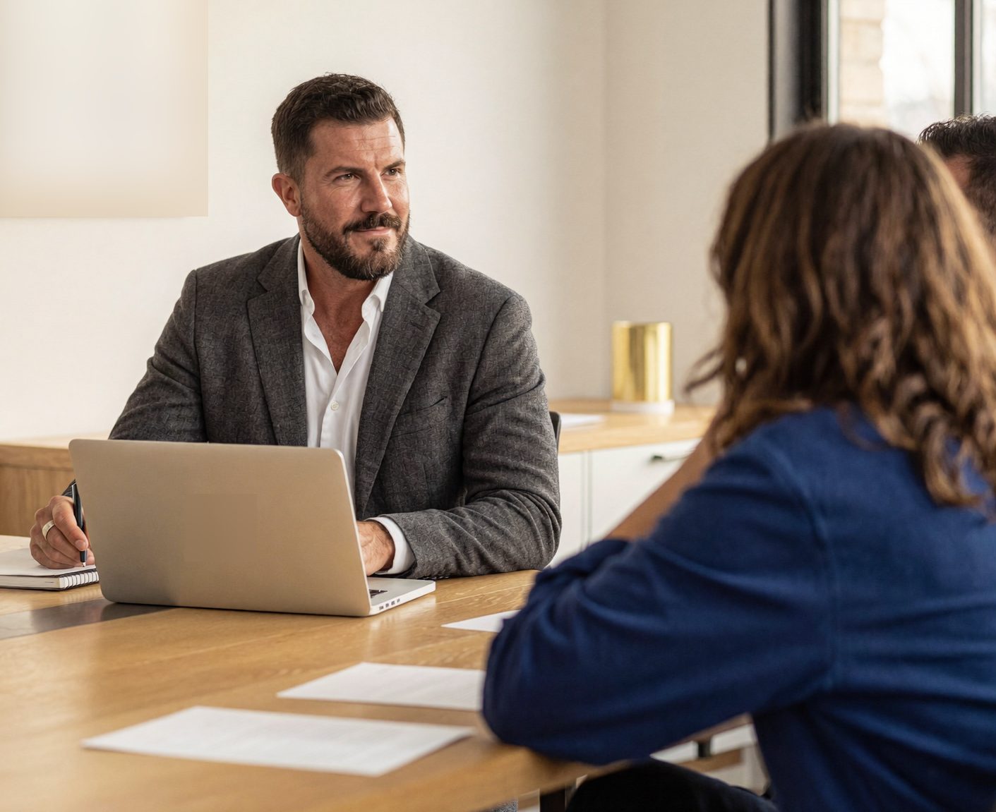 A bearded man in a charcoal blazer leads a working session at a long oak table